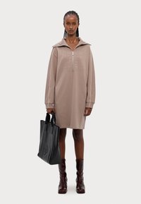 Abito felpa beige con colletto zip, caratterizzato da un logo testurizzato. Abbinato a una borsa tote nera e stivali bordeaux fino al ginocchio.