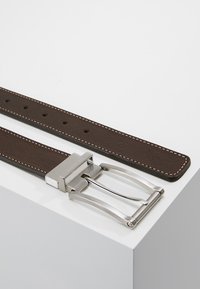 Brauner Leder Gürtel mit weißen Nähten, rechteckiger silberner Metallschnalle und Schlaufe, mit mehreren Einstichlöchern entlang des Riemens.