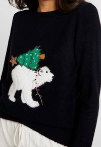 Maglione nero con un orso polare bianco che porta un albero di Natale decorato di verde avvolto in luci colorate.