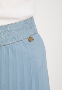 Hellblauer, plissierter Rock mit texturiertem Bund, der glitzernde Akzente aufweist, und einer dekorativen goldenen runden Plakette an der Seite.