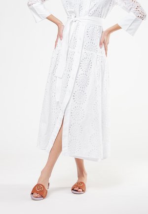 Donna in abito midi bianco con motivo ad occhielli, bottoni e cintura da annodare, indossa sandali marroni aperti con emblema decorativo.