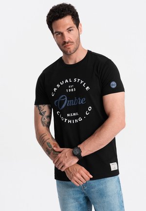 Black cotton t-shirt with white and navy print reading "Casual Style Ombre" and a small logo patch. Short sleeves and relaxed fit.