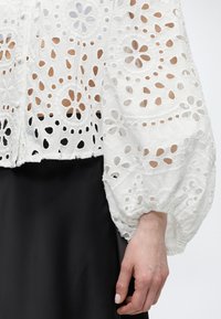 Λευκό πουκάμισο με δαντέλα eyelet και ανθισμένες ανοίγματα, με φουσκωτά ρυτιδωμένα μανίκια, σε συνδυασμό με μαύρη φούστα, που δείχνει ένα χέρι που ακουμπάει στο πλάι.