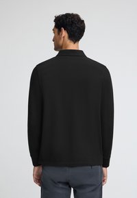 Svart långärmad polo med krage och rak fåll. Slät textur, minimalistisk design och avslappnad passform, sedd bakifrån.