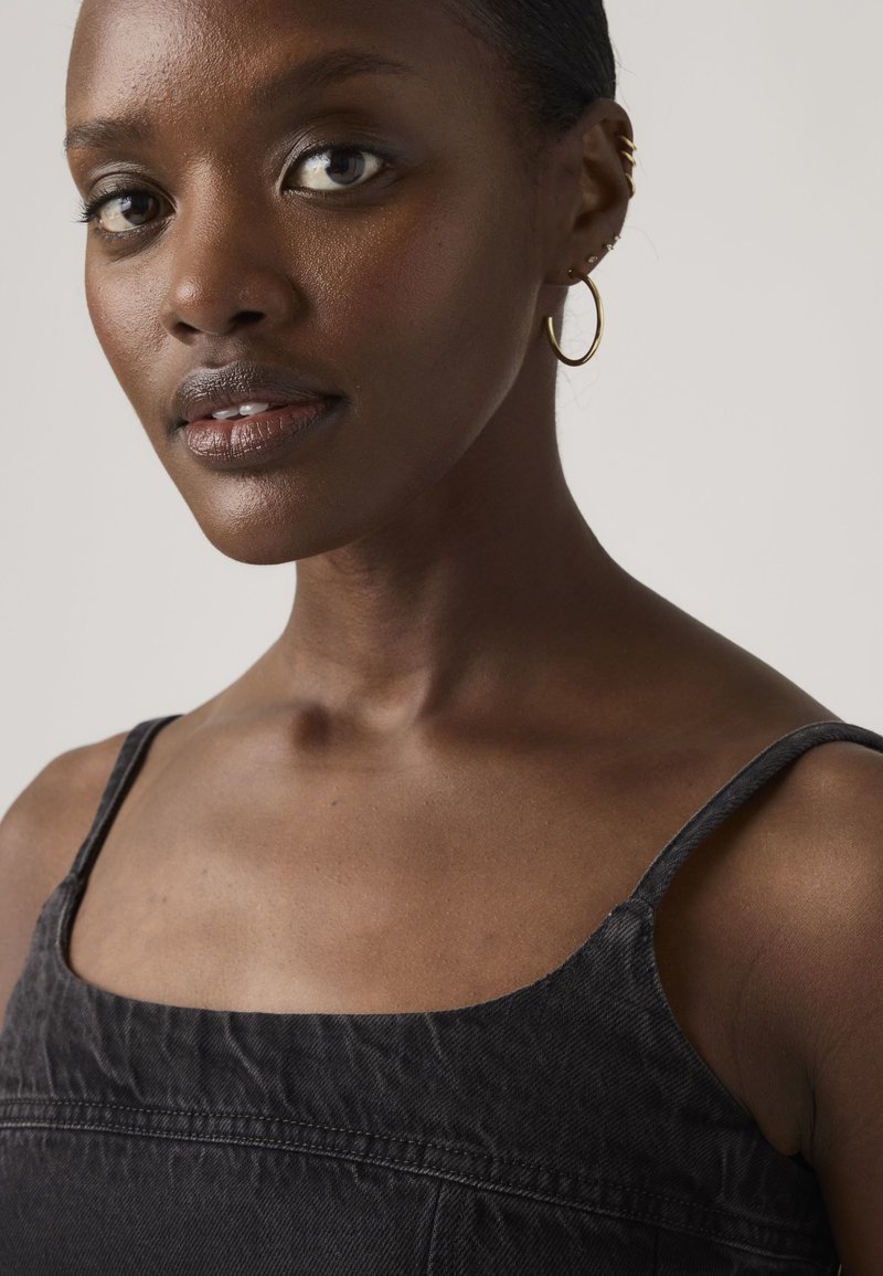 Femme à la peau foncée et aux cheveux courts portant des créoles en or et des bracelets d'oreille, vêtue d'un haut noir sans manches, regardant légèrement sur le côté.