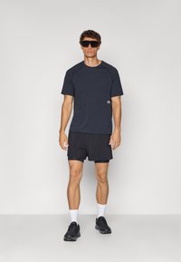 Black athletic t-shirt with short sleeves, paired with black shorts. Model wears dark sunglasses and black sneakers with white socks.