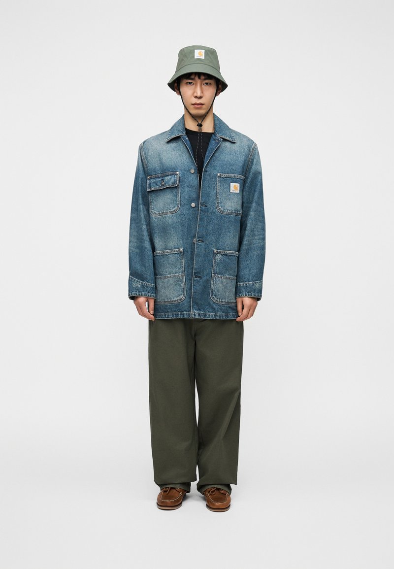 Veste en denim bleu clair avec plusieurs poches, associée à un pantalon vert olive et un chapeau bob gris arborant un logo. Des chaussures marron complètent la tenue.