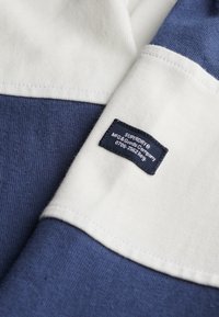 Blauw en wit textiel met een label. Bevat een effen blauwe sectie, witte accenten en een etiket met merk- en verzorgingsinformatie.