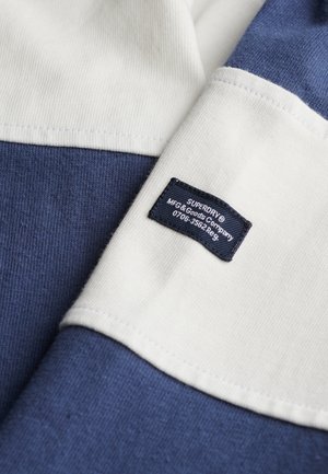 Tessuto testurizzato blu e bianco con un'etichetta. Presenta una sezione blu monocolore, accenti bianchi e un cartellino con informazioni sulla marca e sulle istruzioni per la cura.