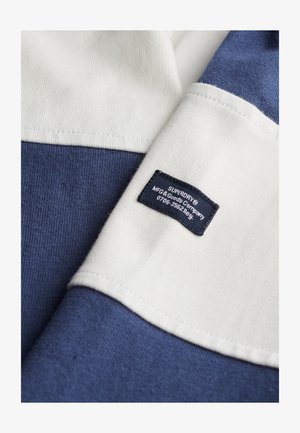 Blå og hvid tekstureret stof med et mærke. Har en solid blå sektion, hvide accenter og et tag med brand- og plejeoplysninger.