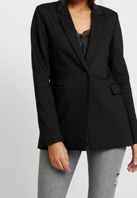 Blazer nero con chiusura a un bottone, rever a punta e due tasche frontali. Tessuto liscio, design sartoriale e vestibilità leggermente strutturata.