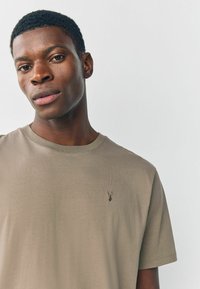 Beige színű pamut póló, kerek nyakkal, rövid ujjakkal és egy kis hímzett logóval a bal mellen. Sima textúrájú.