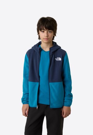 Sudadera con capucha de forro polar en azul marino y turquesa, con cremallera frontal y capucha. Se lleva sobre una camisa color azul verdoso, con detalle de logo en el pecho y las mangas.