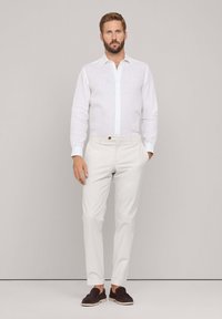 Witte linnen shirt met knoopsluiting, gecombineerd met lichtbeige broeken en donkerbruine loafers, staand tegen een neutrale achtergrond.