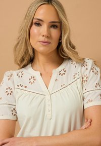Ivory top with floral eyelet detailing on sleeves, button placket, and a relaxed fit. Soft texture and light, breathable fabric.