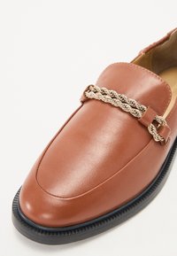 Brūni ādas loafers ar vērptu metāla ķēdes akcentu un teksturētu gumijas zoli. Gluda virsma un elegants dizains.