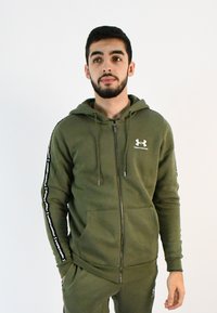 Sudadera con cremallera en verde oliva, con capucha, bolsillos frontales y franjas laterales negras que presentan el nombre de la marca. Fabricada con un tejido suave y texturizado.