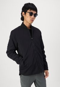 Black bomber jacket with a zip closure, ribbed collar, and side pockets; worn with dark sunglasses and gray pants on a plain background.