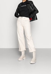 Jeans bianchi a vita alta con orli arrotolati, abbinati a una maglietta nera e una giacca di pelle nera, completati da stivali color crema.