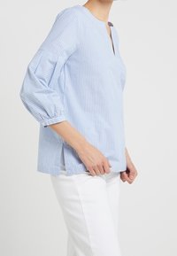 Blouse rayée bleu clair avec un col à patte, des manches bouffantes trois-quarts et des fentes sur les côtés, fabriquée en tissu doux. Associée à un pantalon blanc.