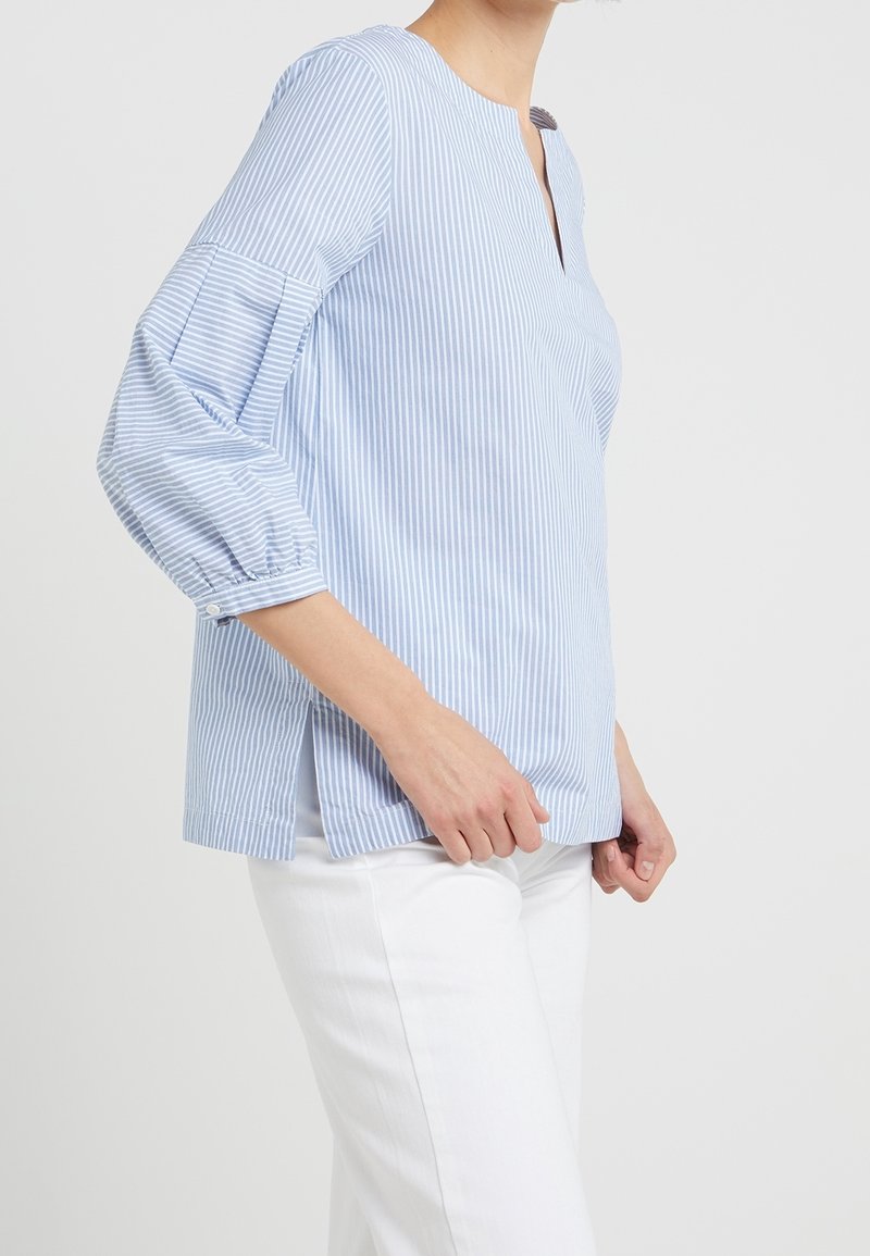 Blouse rayée bleu clair avec un col à patte, des manches bouffantes trois-quarts et des fentes sur les côtés, fabriquée en tissu doux. Associée à un pantalon blanc.