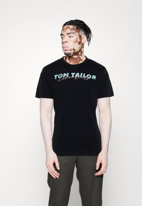 T-shirt en coton noir avec "TOM TAILOR Essentials" imprimé en turquoise et rose. Manches courtes et col rond. Texture lisse.