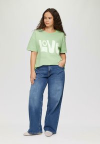 Jasnozielona bawełniana koszulka z napisem "LOVE" w kolorze białym, połączona z niebieskimi szerokimi dżinsami. Model stoi z jedną ręką w kieszeni.