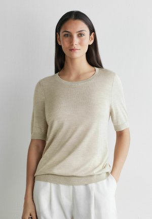 Frau mit langem dunklem Haar, die ein beiges Strickoberteil mit kurzen Ärmeln und eine weiße Hose trägt, steht vor einer schlichten hellen Wand.