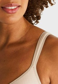 Close-up van het onderste gezicht van een persoon, hals en schouder die een beige textuur tanktop draagt met krullend haar zichtbaar op een witte achtergrond.