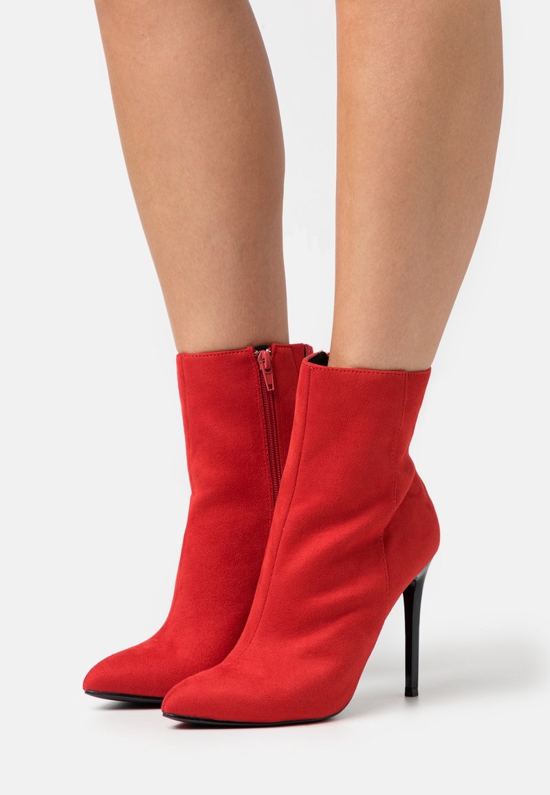 Paire de bottines en daim rouge avec bouts pointus, fermeture éclair latérale et talons aiguilles noirs portées sur des jambes nues.