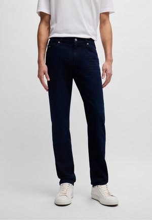 Hombre vestido con pantalones vaqueros ajustados de color azul oscuro, camisa blanca de malla de manga corta y zapatillas blancas, de pie sobre un fondo liso.