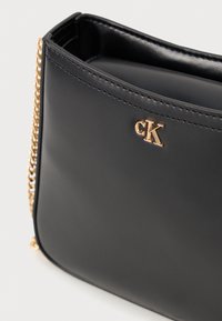 Čierna kožená kabelka s hladkou textúrou, s zlatou kovovou reťazou a akcentom loga. Dizajn s otvoreným vrchom umožňuje ľahký prístup.