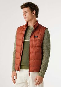Gilet imbottito color ruggine con colletto alto e zip frontale, indossato sopra un maglione verde testurizzato. Presenta un motivo a costo orizzontale.