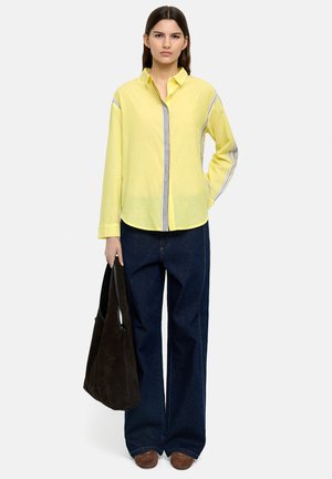 Młoda kobieta w żółtej bluzce z długim rękawem, ciemnych szerokich dżinsach, brązowych butach, trzymająca dużą czarną torbę na ramię, stojąca na białym tle.