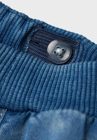 Cintura de denim azul con textura acanalada, asegurada con un botón de plástico y una tela oscura de contraste debajo.