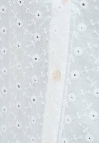 Weiße gestickte Bluse mit floralen Mustern, Knopfleiste und transparenter Stofftextur; zarte Stickdetails sind sichtbar.