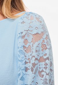 Kvinne iført lyseblå topp med gjennomsiktig blomstermønstret blonder erme som viser detaljert blonde mønster og huden under.