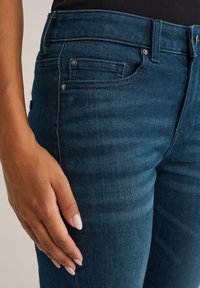 Temno modri denim jeans z ozkim krojem, sprednjimi in zadnjimi žepe ter diskretnimi šivi. Roka resting na stegnu.