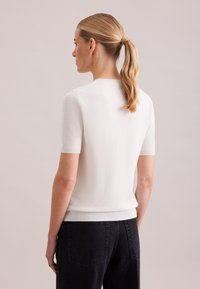 Maglione bianco a maniche corte in maglia con scollo rotondo e orlo a coste, abbinato a jeans neri. Texture liscia senza motivi visibili.