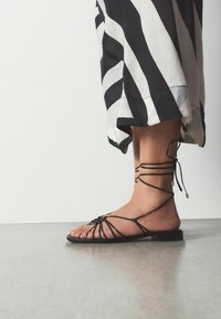 Pied nu dans une sandale plate noire à bretelles avec des lacets autour de la cheville, à côté d'une jupe ample à rayures noires et blanches, debout sur un sol gris.