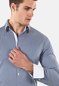 Camicia a quadretti blu e bianchi con colletto a punta, polsini bianchi e tessuto sottile; bottoni visibili nella parte anteriore.