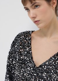 Top en sequins noirs avec un décolleté en V profond et des manches courtes, mettant en valeur des sequins ronds brillants sur un tissu texturé.