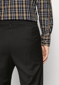Zwarte broek met een gladde textuur, met een achterzak. Vergezeld van een fitted, geruite shirt in zwart, bruin en witte tinten.