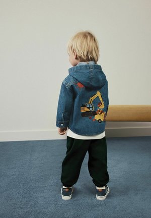 Junges Kind mit blondem Haar steht mit dem Rücken zur Kamera und trägt eine blaue Jeansjacke mit einer Stickerei eines Baumaschinenfahrzeugs auf dem Rücken.