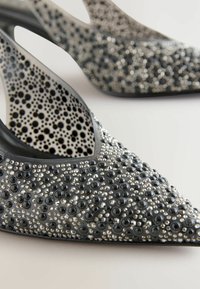 Schwarze High Heels mit spitzer Zehenpartie, die eine strukturierte Oberfläche aufweisen, die mit verschiedenen Größen von schwarzen und silbernen Verzierungen geschmückt ist.