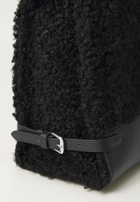 Black bag featuring a textured, curly wool exterior with a smooth leather base and a detachable leather strap secured by a silver buckle.