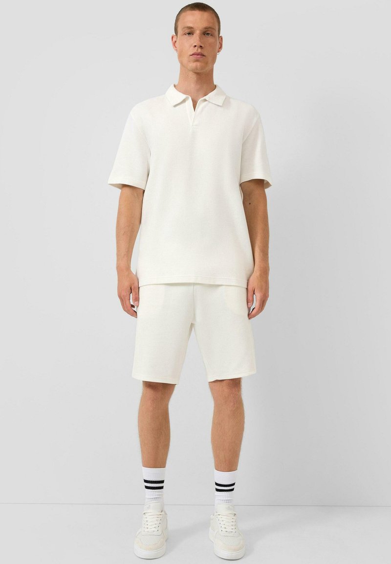 Jeune homme debout portant un polo blanc à manches courtes, un short blanc, des baskets blanches et des chaussettes blanches avec deux rayures noires.