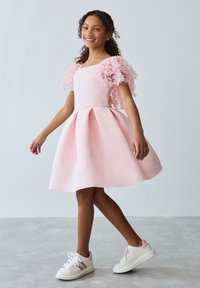 Hymyilevä tyttö vaaleanpunaisessa mekossa, jossa on kukkakuviolliset pöhöhihaiset ja valkoiset tennarit, kävelee minimalistisessa huoneessa, jossa on vaaleanharmaa lattia ja valkoinen seinä.