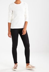 White knitted sweater with long sleeves, paired with black skinny jeans and metallic rose gold slip-on sneakers. Fitted design, mid-calf length.