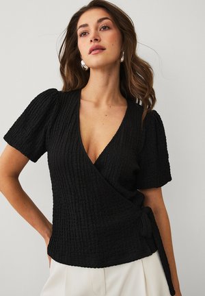 Kvinde med løst bølget hår iført sølv hoop øreringe og en tekstureret sort omslagstop med puffede ærmer og hvide bukser.